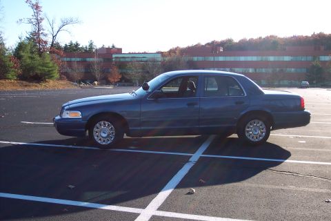 2001 Ford Crown Victoria - A blue 2001 Ford Crown Victoria is parked in a lot with a building in the background.