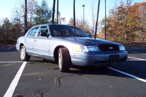 2001 Ford Crown Victoria - A blue 2001 Ford Crown Victoria is parked in a lot with trees in the background.