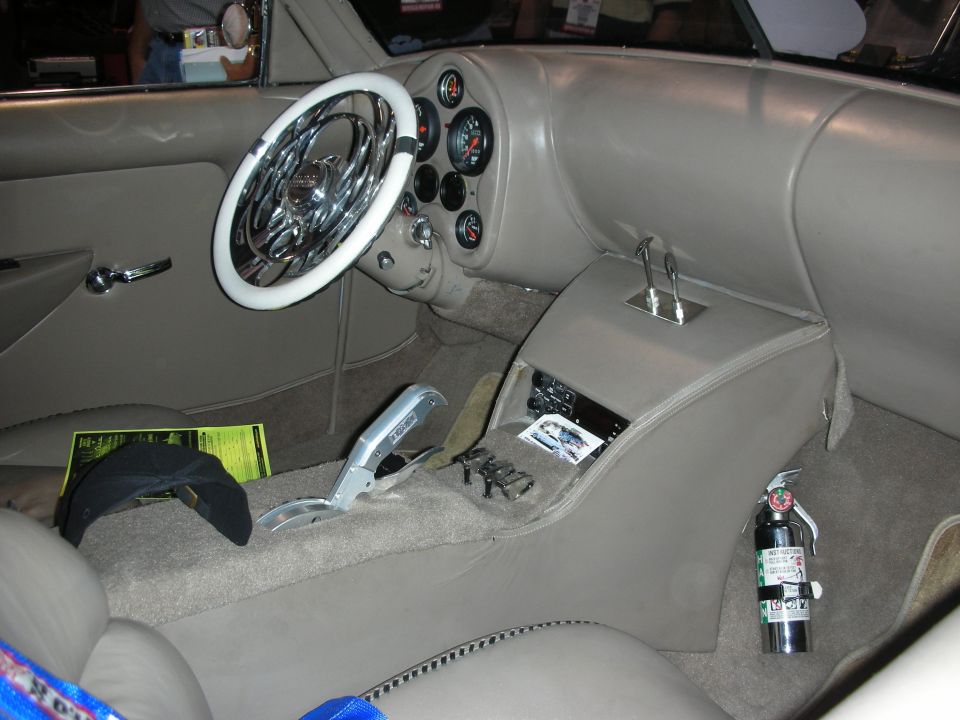 Custom car interior with a white steering wheel, dashboard gauges, and a chrome shifter. A fire extinguisher is visible on the right