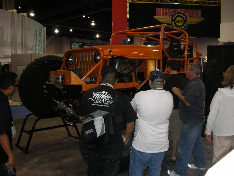 Orange off-road vehicle with large tires and roll cage, viewed from the front at an indoor event