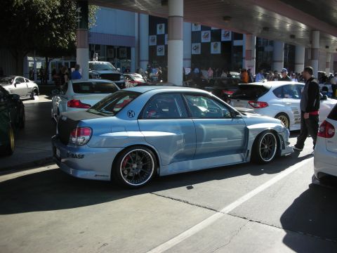 1998 Ford Crown Victoria - A modified Subaru WRX STI is parked in a lot with other vehicles and people milling around.