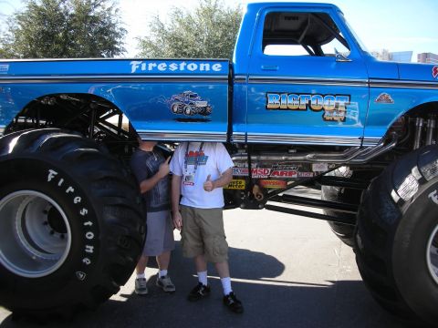 1998 Ford Crown Victoria - A blue Bigfoot monster truck is shown with two people standing underneath it.