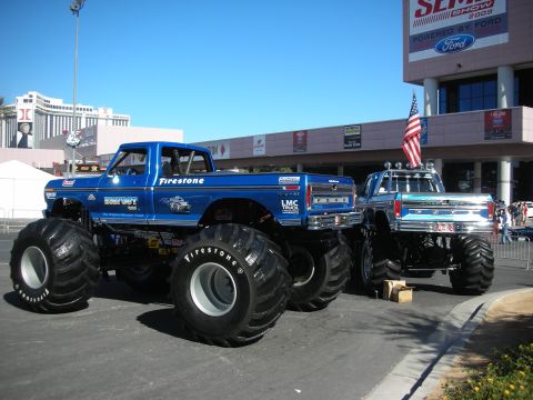 1998 Ford Crown Victoria - Two monster trucks, one blue with 'Bigfoot' branding and another chrome-accented, are parked outdoors.