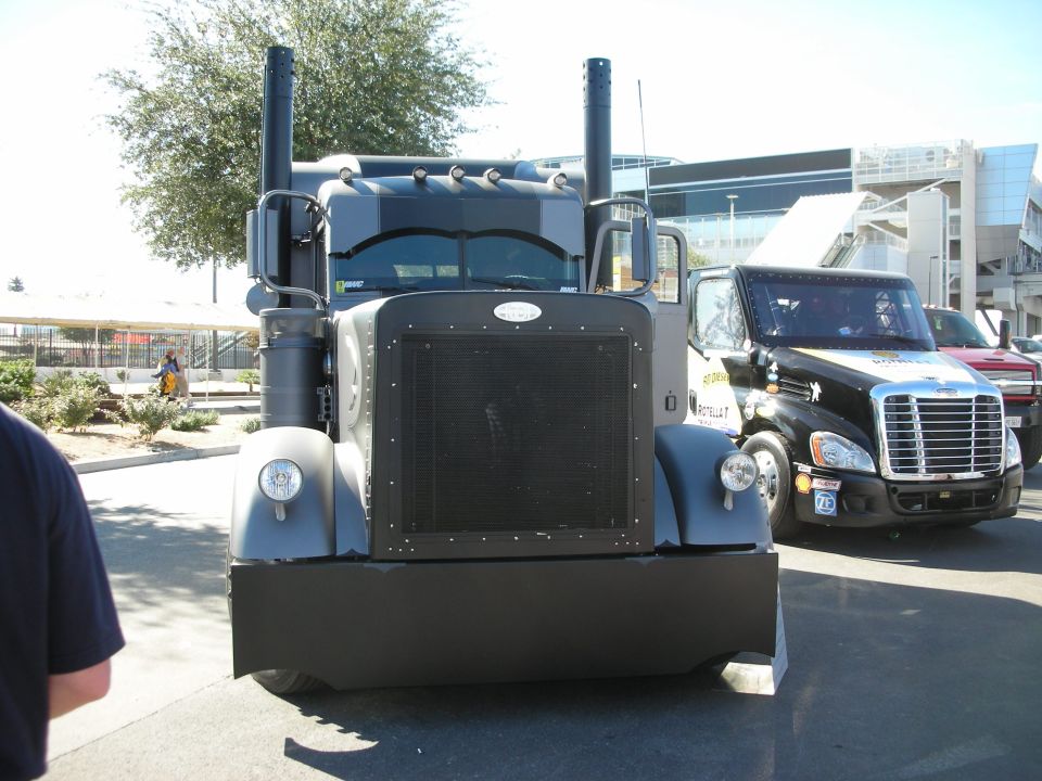 Front view of a custom grey semi-truck with large exhaust stacks, parked next to a black semi-truck with racing decals