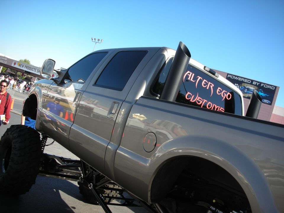 Gray lifted pickup truck with large exhaust pipes and 'Alter Ego Customs' on the rear window
