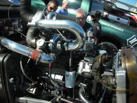 1998 Ford Crown Victoria - A highly customized engine with a prominent turbocharger and polished intake piping is examined by onlookers, with a green truck in the bac…