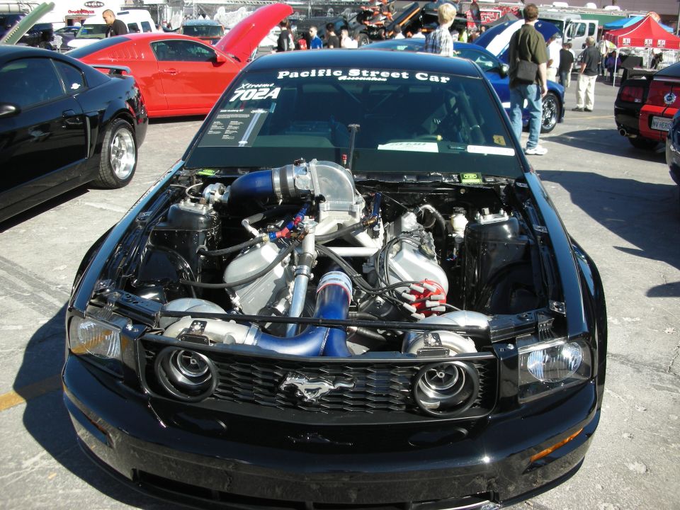black Ford Mustang with engine exposed, twin turbos, and custom intake. Pacific Street Car 702A sticker on windshield. Red Mustang and black car in background