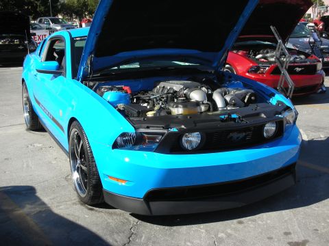 1998 Ford Crown Victoria - A bright blue Ford Mustang is parked with its hood open, revealing the engine compartment. Another red Mustang is visible in the background.