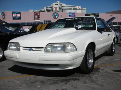 1998 Ford Crown Victoria - A white Ford Mustang is parked outside a building with a 'Hot Rod' sign, showing its front end and custom hood.
