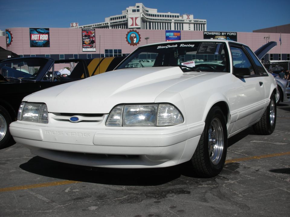 White Ford Mustang with a scoop hood and racing decals on the windshield. Another car is parked next to it