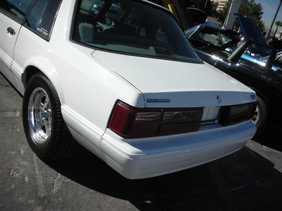 White police interceptor sedan rear, chrome wheels, dark tinted windows, parked next to black car with hood up
