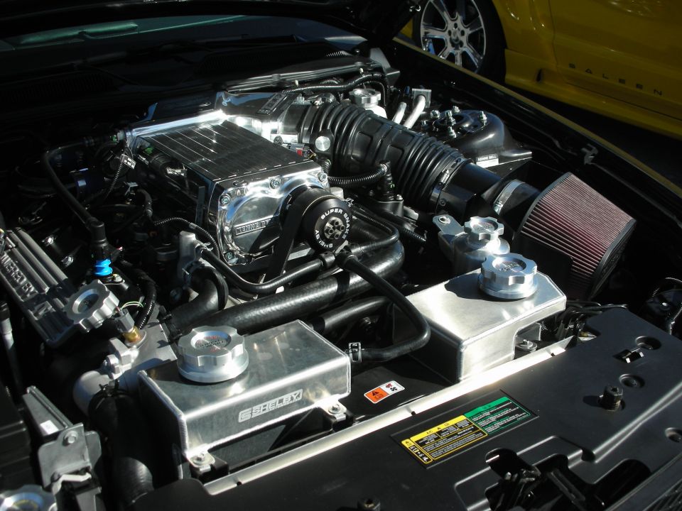 Engine bay with chrome supercharger, Shelby branded components, and a yellow Saleen Mustang in the background