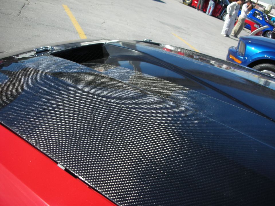 close-up of a car's carbon fiber hood with a vent, other cars and people in background