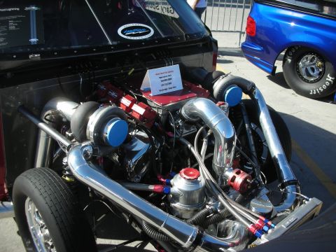 1998 Ford Crown Victoria - Custom twin-turbocharged V8 engine on display, with a blue pickup truck in the background.