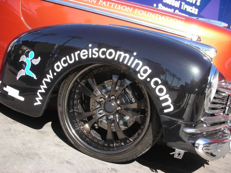 Black fender with www.acureiscoming.com text and large chrome wheels. Orange car behind