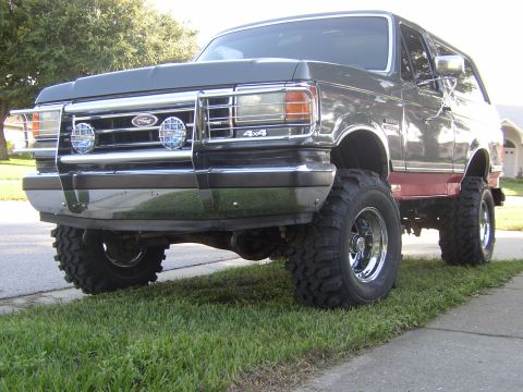 1991 Ford Bronco - Low front three-quarter shot of a lifted gray Ford Bronco with a maroon lower stripe parked over a sidewalk in a suburban yard. It wears a…