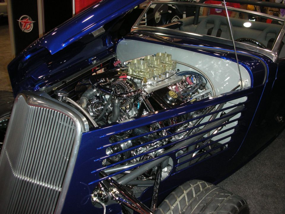 Blue hot rod with hood open showing a V8 engine with multiple carburetors. Front grille is chrome