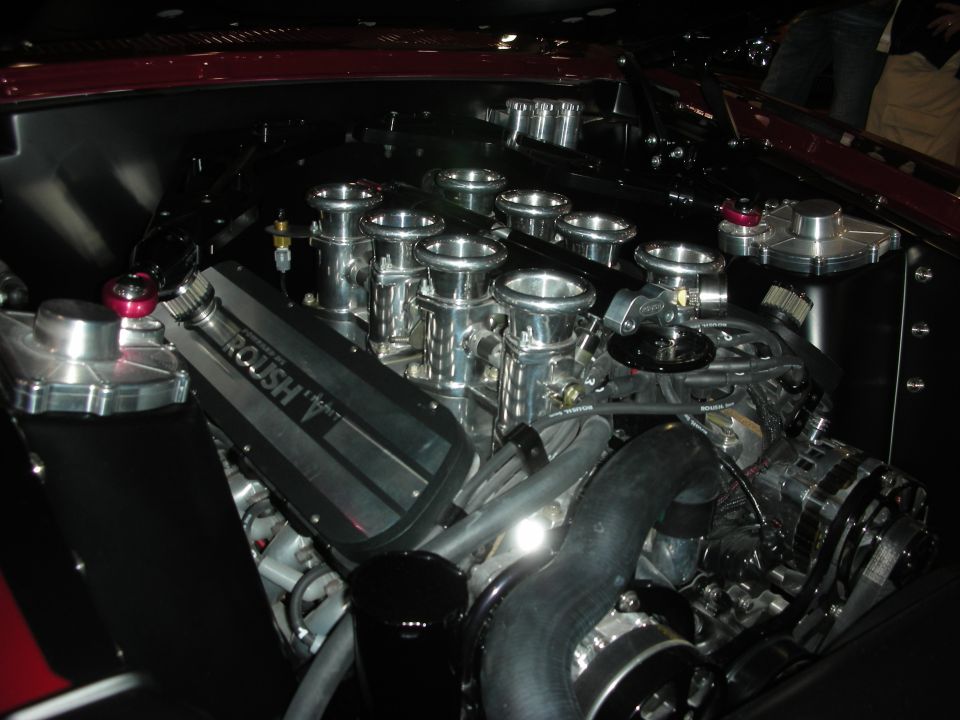 Engine with "ROUSH" branding on a black intake manifold, chrome velocity stacks, and a black alternator
