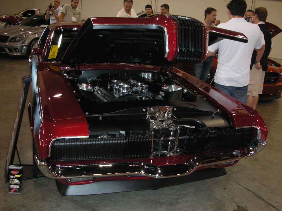 Customized classic car with hood open showing engine, front grille visible. Other cars in background