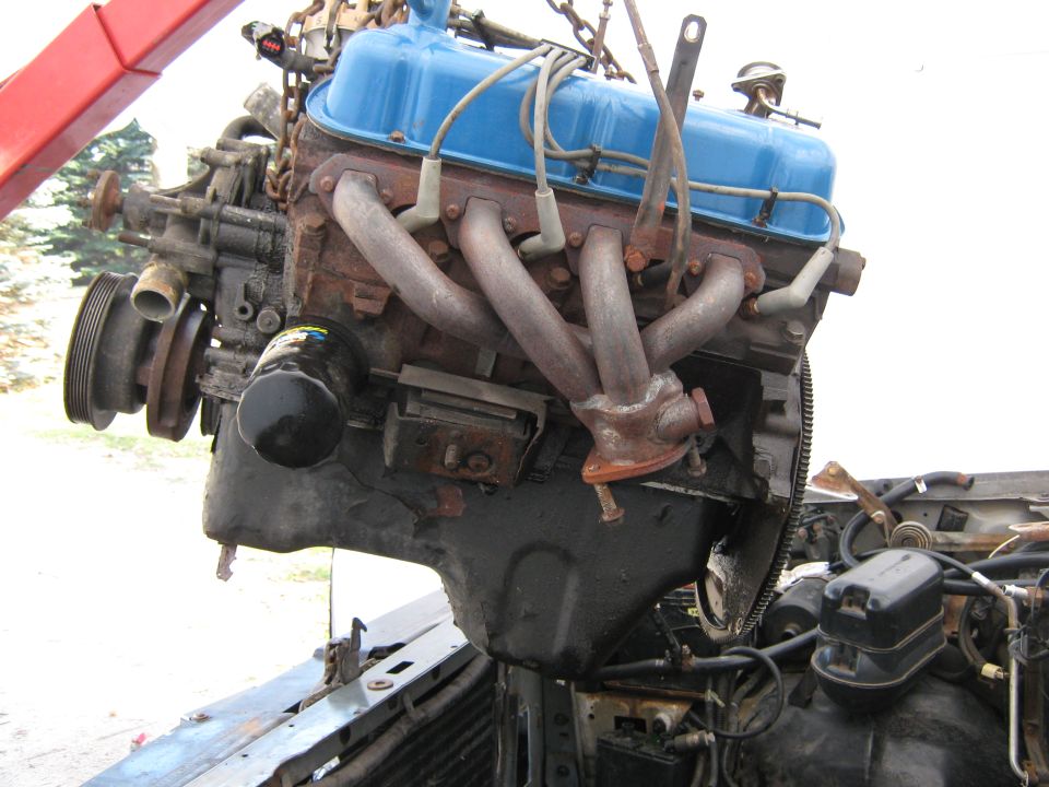 Engine with blue valve covers and rusted exhaust manifolds being lifted by a red crane