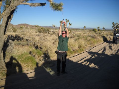 1986 Ford Bronco II - A man holds a bottle aloft in a desert scene, with a white SUV visible in the background on a sandy track.