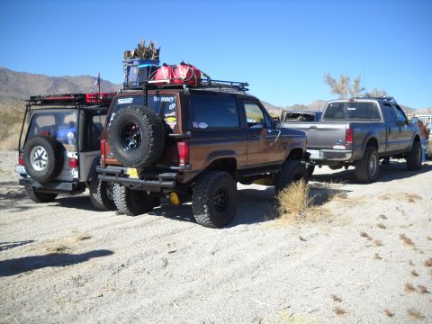 1986 Ford Bronco II - Multiple modified off-road vehicles parked in a sandy, arid landscape under a clear blue sky.