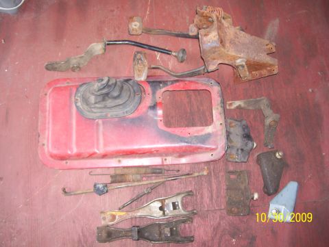 1978 Ford Bronco - A collection of assorted, aged vehicle parts, including pedals and transmission components, are displayed on a red surface.