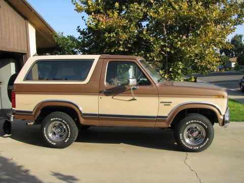 1986 Ford Bronco - This two-tone Ford Bronco is parked outside a garage with trees in the background.