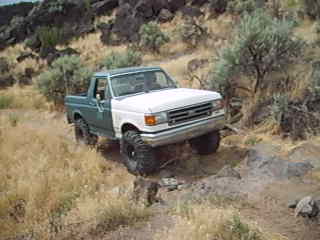 1989 Ford Bronco - A two-tone Ford Bronco navigates a rocky, off-road path in a dry, arid landscape.