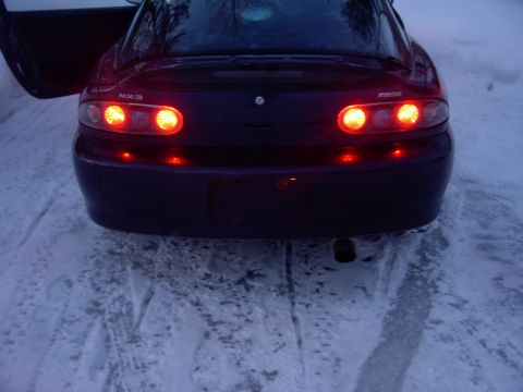1990 Ford Bronco - The rear of a dark Mazda MX-3 is visible with its taillights illuminated, parked in a snowy environment.