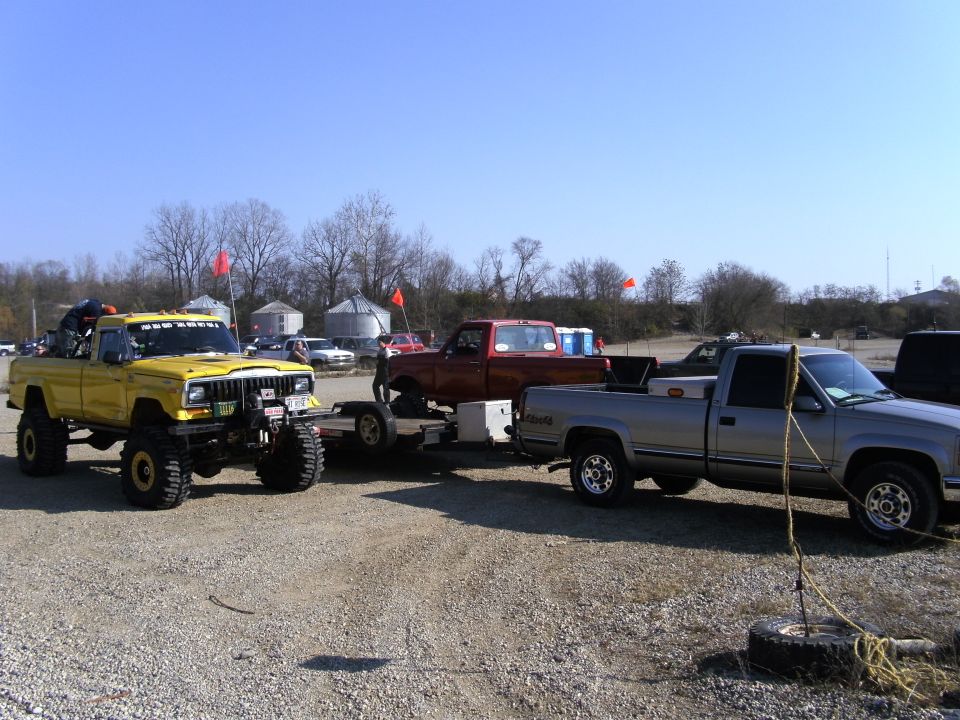 Yellow Jeep Comanche pickup with big tires, towing a trailer. A red truck and a silver truck are behind it