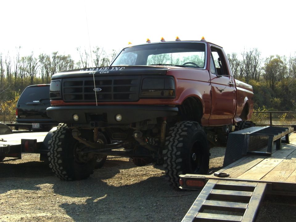 Red lifted Ford pickup truck with black grille and hood decal, parked on gravel next to a trailer