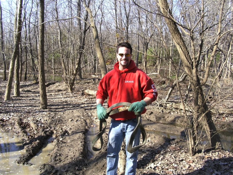 Man in red hoodie and green gloves holds a tow strap in a muddy, wooded area. Watermark reads SUPERMOTORS.NET