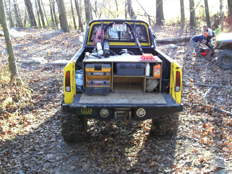 Yellow pickup truck bed loaded with tools and equipment, with people working near another vehicle in the background