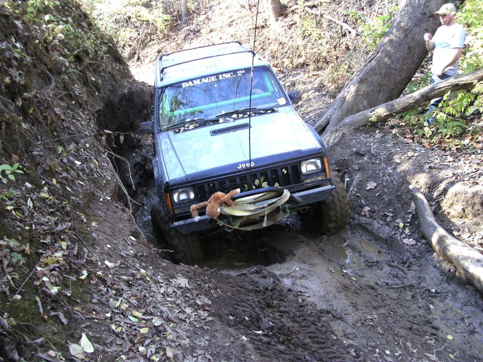 Jeep Cherokee XJ navigating a muddy ditch with a fallen tree nearby. 'DAMAGE INC.' text on windshield