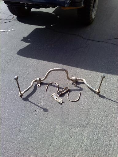 1996 Ford Bronco - Anti-roll bar and associated hardware laid out on a paved surface, with the rear tire and fender of a vehicle visible in the background.