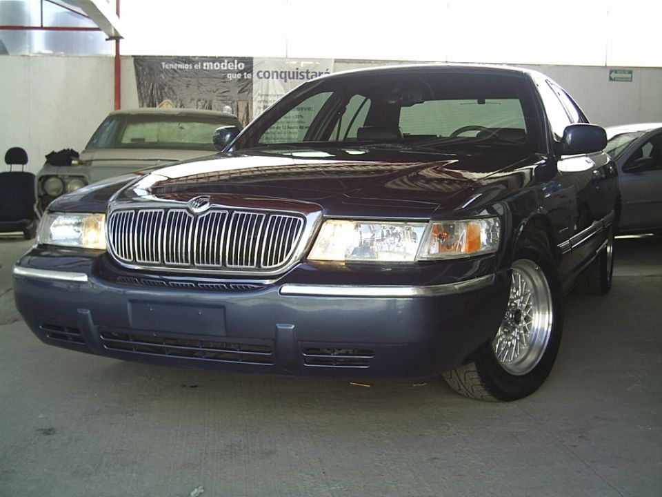 Dark colored Mercury Grand Marquis front end with chrome grille and aftermarket wheels. Another car is visible behind it