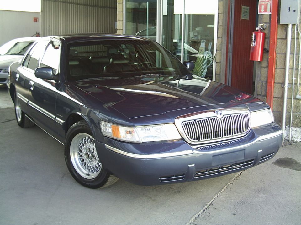 Navy blue Mercury Grand Marquis sedan, front 3/4 view, chrome wire wheels, outside shop