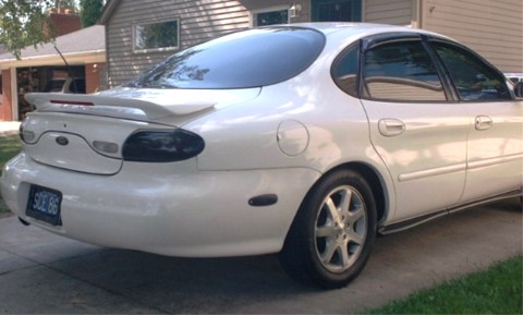 1990 Ford Taurus - This white sedan has a spoiler and tinted windows, parked in front of a house.