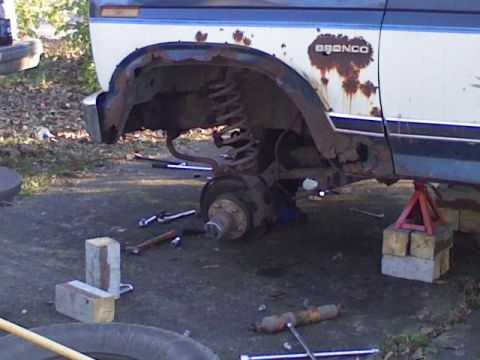 1986 Ford Bronco - A heavily rusted Ford Bronco is shown with its front wheel removed and the vehicle elevated on jack stands.