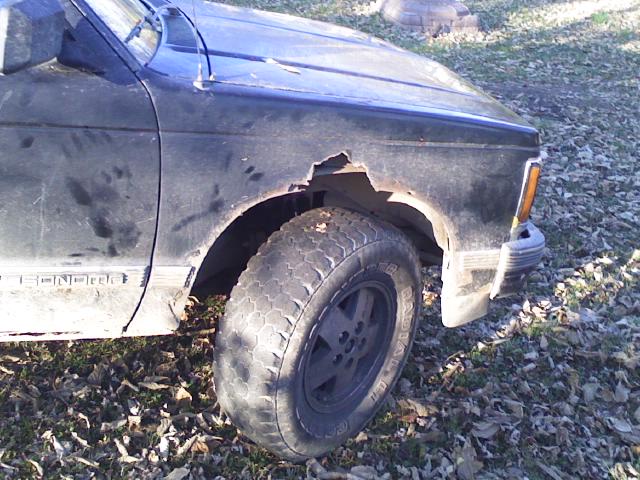 Rusted-out front fender of a black compact pickup over an all-terrain tire on leafy ground