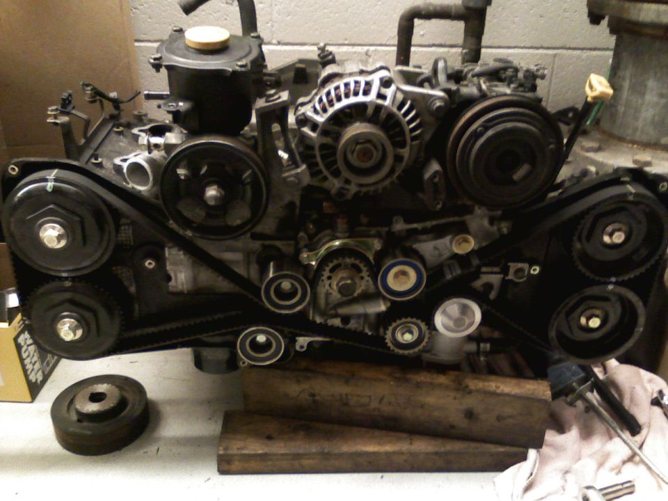 Subaru boxer engine on a bench with timing covers off exposing belt and cam sprockets
