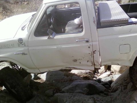 1981 Ford F-150 - White pickup truck with significant body damage on the passenger side, off-roading over rocks.