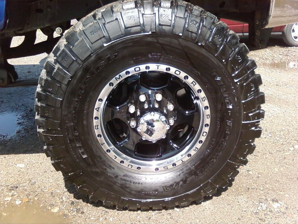 Truck tire with aggressive tread and black wheel with chrome center cap. A blue truck frame is visible above