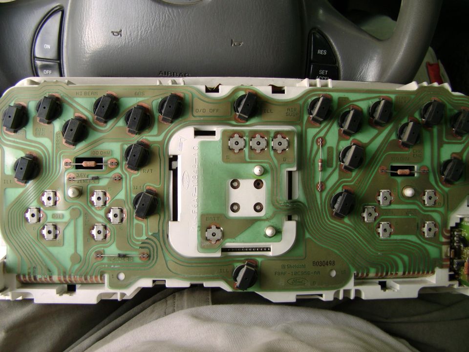 Instrument cluster circuit board with black knobs and green traces, partially visible steering wheel and dashboard above