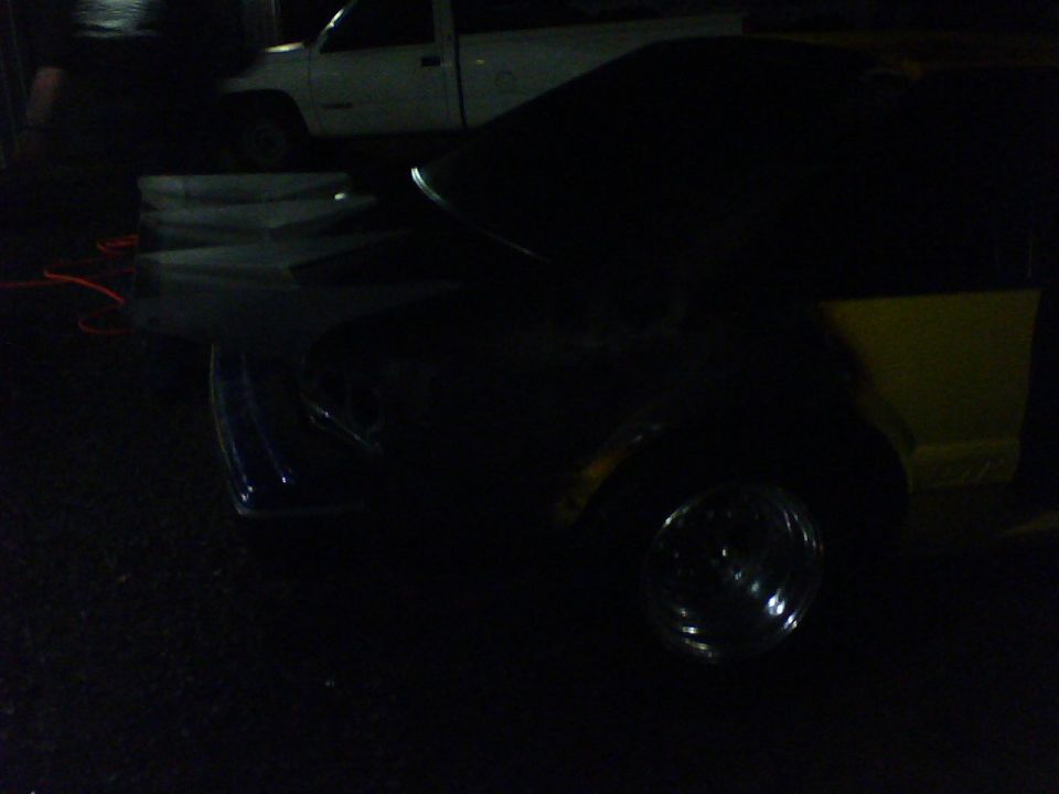 Dark car front quarter view, yellow rear quarter panel, chrome wheel, white truck in background