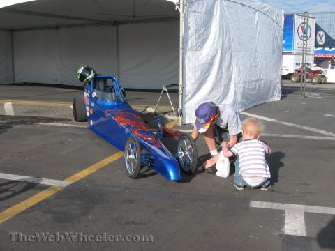1968 Ford Mustang - A man and a child are near a blue dragster with flame graphics under a large tent.