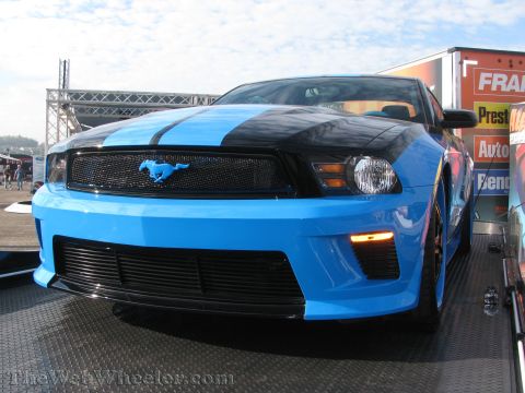 1968 Ford Mustang - A blue and black Ford Mustang with racing stripes is parked on a trailer, with a trailer advertising "Fran's Auto" in the background.