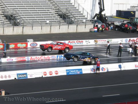 1968 Ford Mustang - Two drag cars, a red Mustang and a blue car with flames, launch off the starting line during a race.