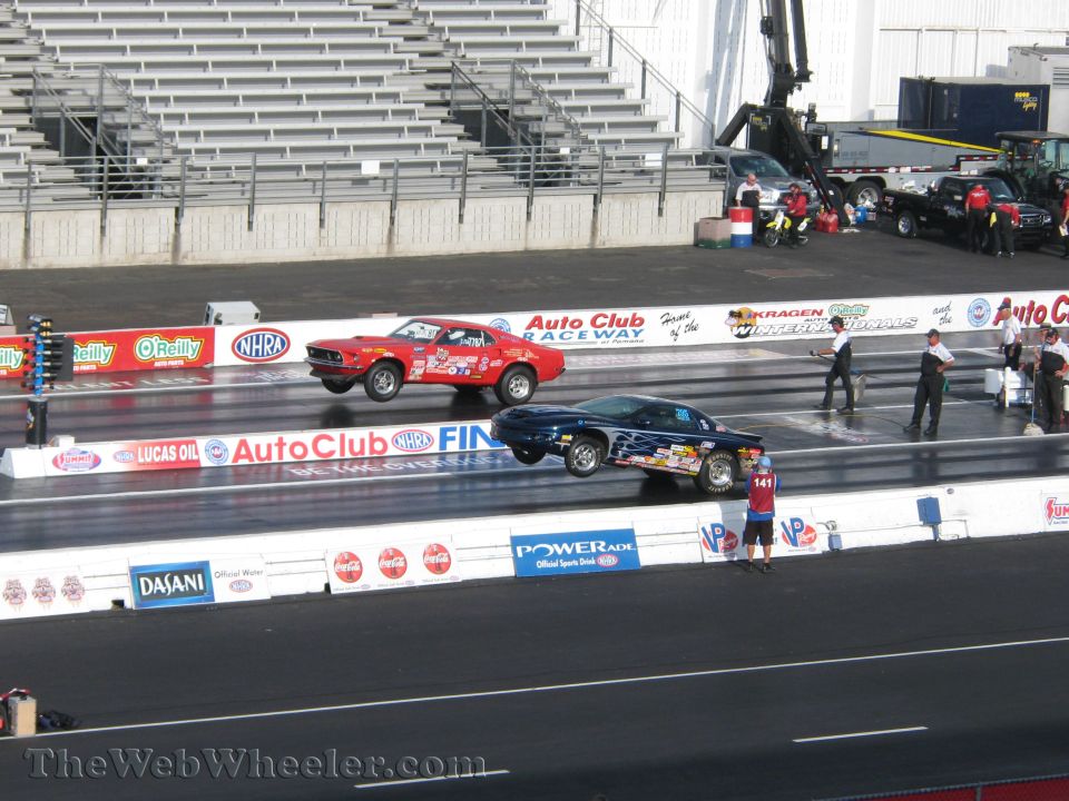 Red 1969 Ford Mustang drag car in mid-air, blue car with flames beside it on track